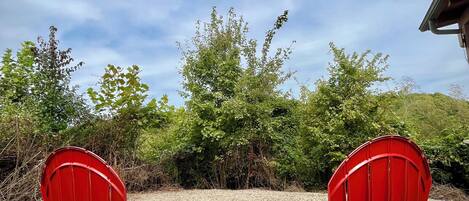 Two red Adirondack chairs facing each other with a gravel ground and greenery in the background.
