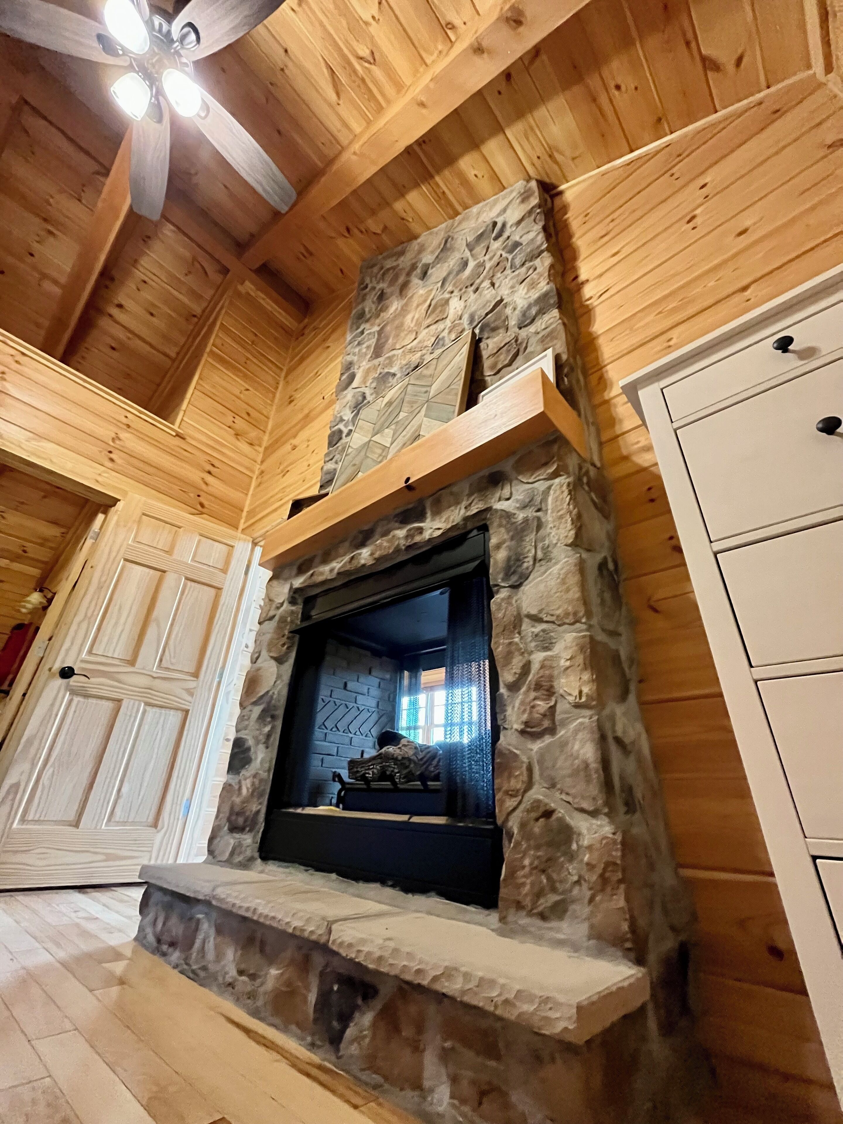 A stone fireplace with a wooden mantel, a ceiling fan, and wooden paneling.