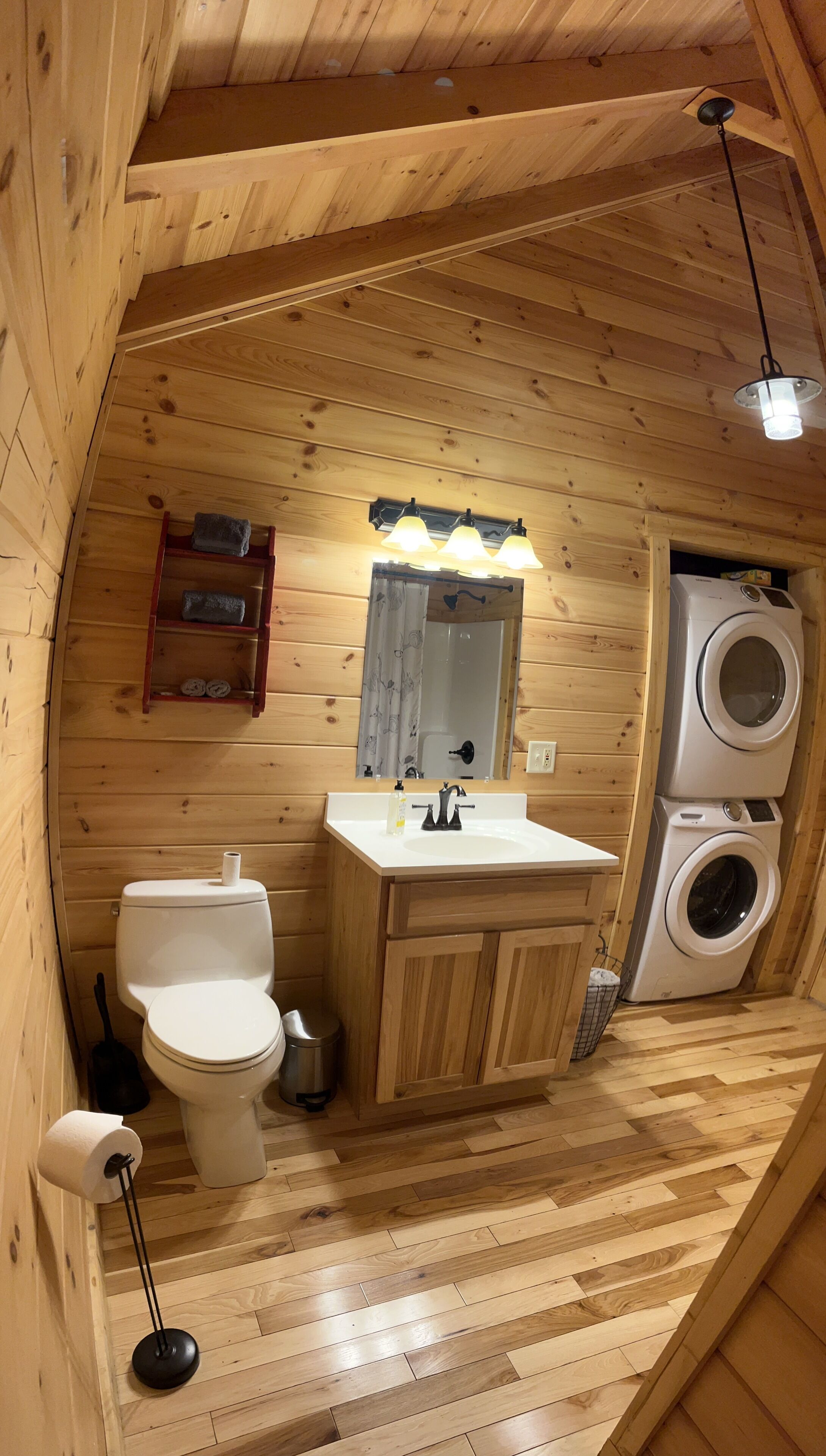 A compact wooden cabin bathroom with a toilet, sink, and stacked washer-dryer.