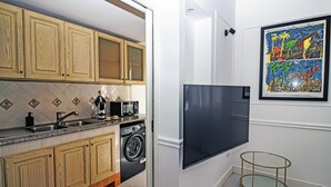 Une cuisine moderne avec des meubles en bois, un lave-linge encastré, un grand téléviseur noir et un tableau abstrait aux couleurs vives accroché au mur.