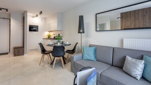 Uma sala de estar moderna com um sofá cinzento, uma mesa de jantar com cadeiras, e uma cozinha ao fundo.