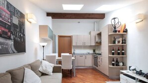 Un espacio habitable compacto con sofá, mesa de comedor, sillas, kitchenette y una estantería para vinos.