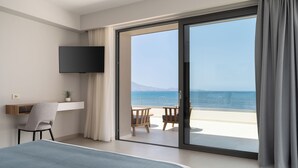 Ein Hotelzimmer mit einem Bett, einem Schreibtisch, einem Sessel, einem Fernseher und einer Glastür zum Balkon mit Meerblick.