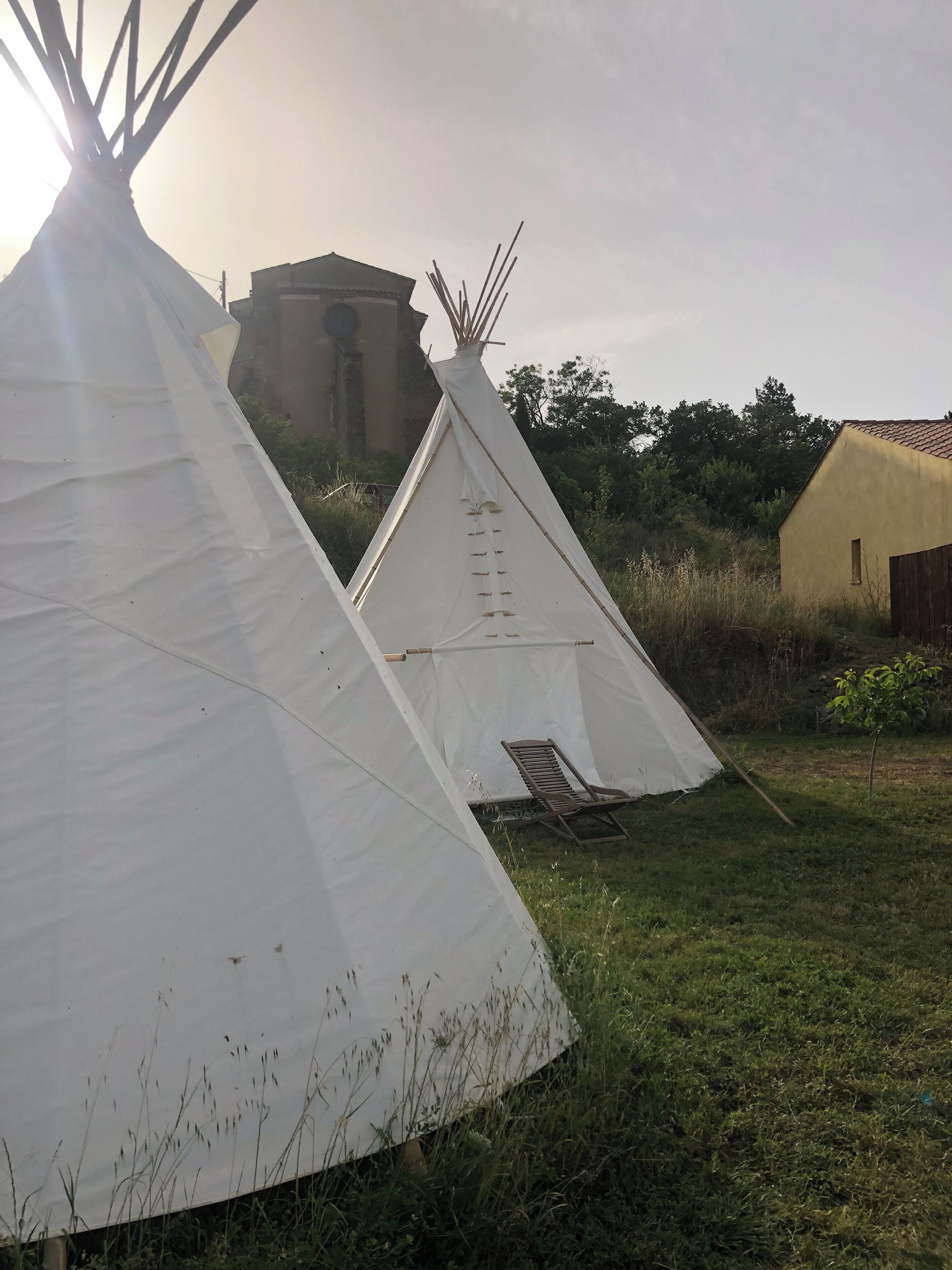 Een witte tipi met een houten stoel van binnen, gelegen in een grasrijk gebied met een geel gebouw en een windmolen op de achtergrond.