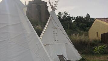 Een witte tipi met een houten stoel van binnen, gelegen in een grasrijk gebied met een geel gebouw en een windmolen op de achtergrond.