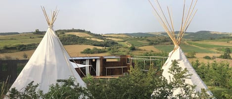 Two white teepees with wooden structures in the background, surrounded by greenery and hills.