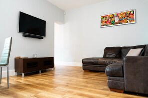 A living room with a flat-screen TV mounted on the wall, a brown leather sofa, a wooden entertainment unit, and a wooden floor.