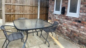 Un patio con un tavolo in vetro e quattro sedie, una recinzione in legno e un muro di mattoni con due finestre.