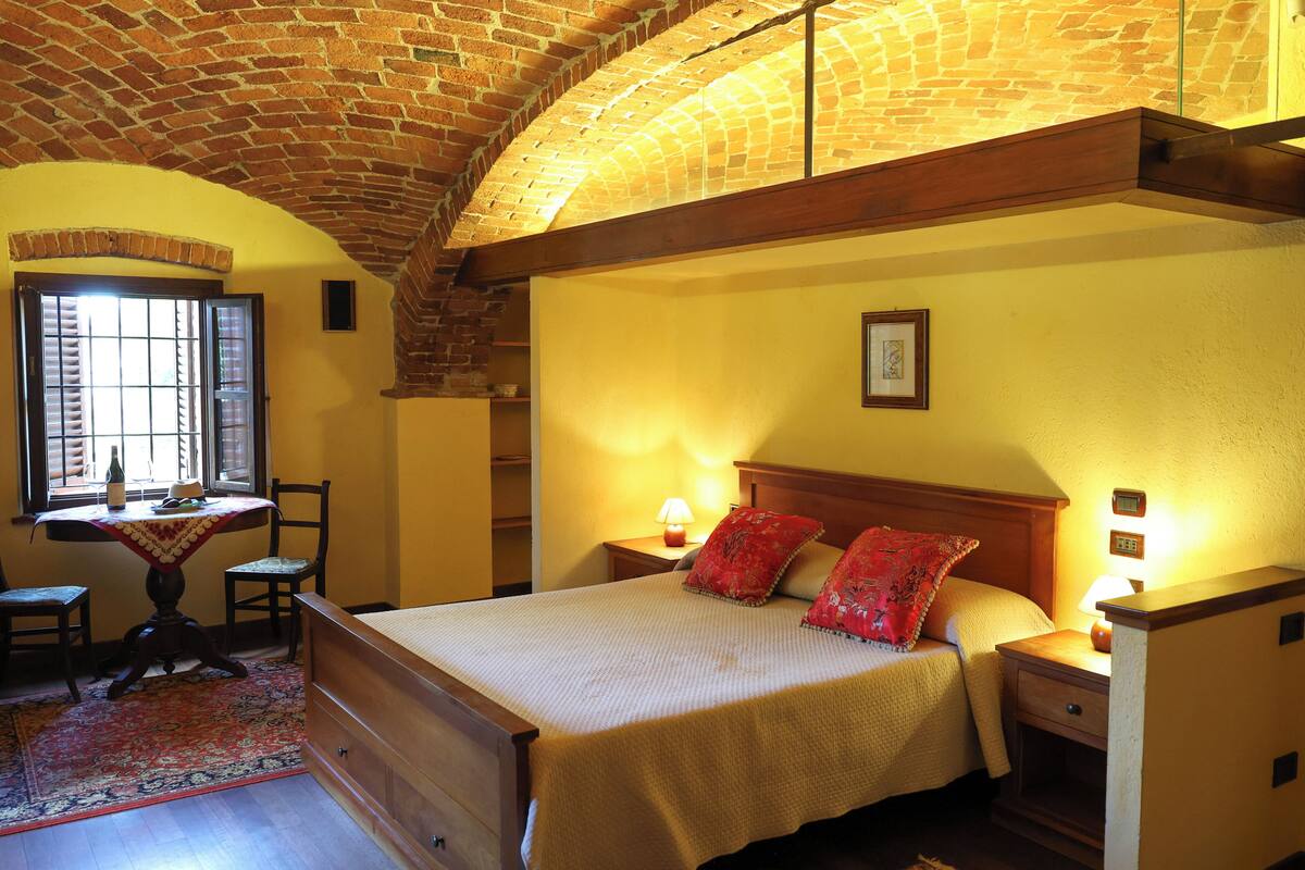 A bedroom with a bed, bedside tables, a nightstand, a window with blinds, a table with chairs, and a patterned rug.