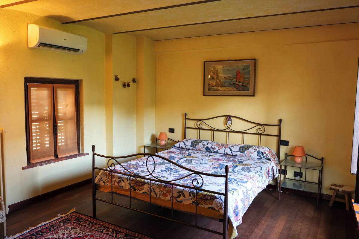 A bedroom with a metal bed frame, a wooden floor, a window with blinds, a wall-mounted air conditioner, a framed painting, and two bedside tables with lamps.