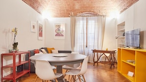 Een eetgedeelte met een bakstenen plafond, een ronde tafel, witte stoelen, een bank en een televisie op een geel schap.