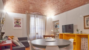 Een moderne woonkamer met een bakstenen plafond, een ronde eettafel, een grijze bank, witte stoelen, een geel boekenkast en een televisie.