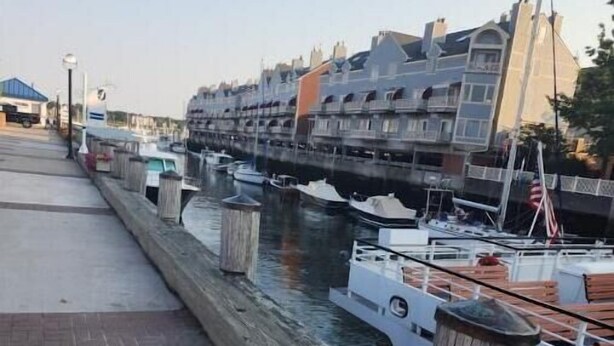 A waterfront promenade with a row of boats docked along the water's edge, wooden bollards separating the walkway from the water, and a row of buildings in the background.