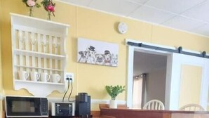 A compact kitchen with a microwave, coffee maker, and small refrigerator. There is a white shelf with cups and a potted plant.