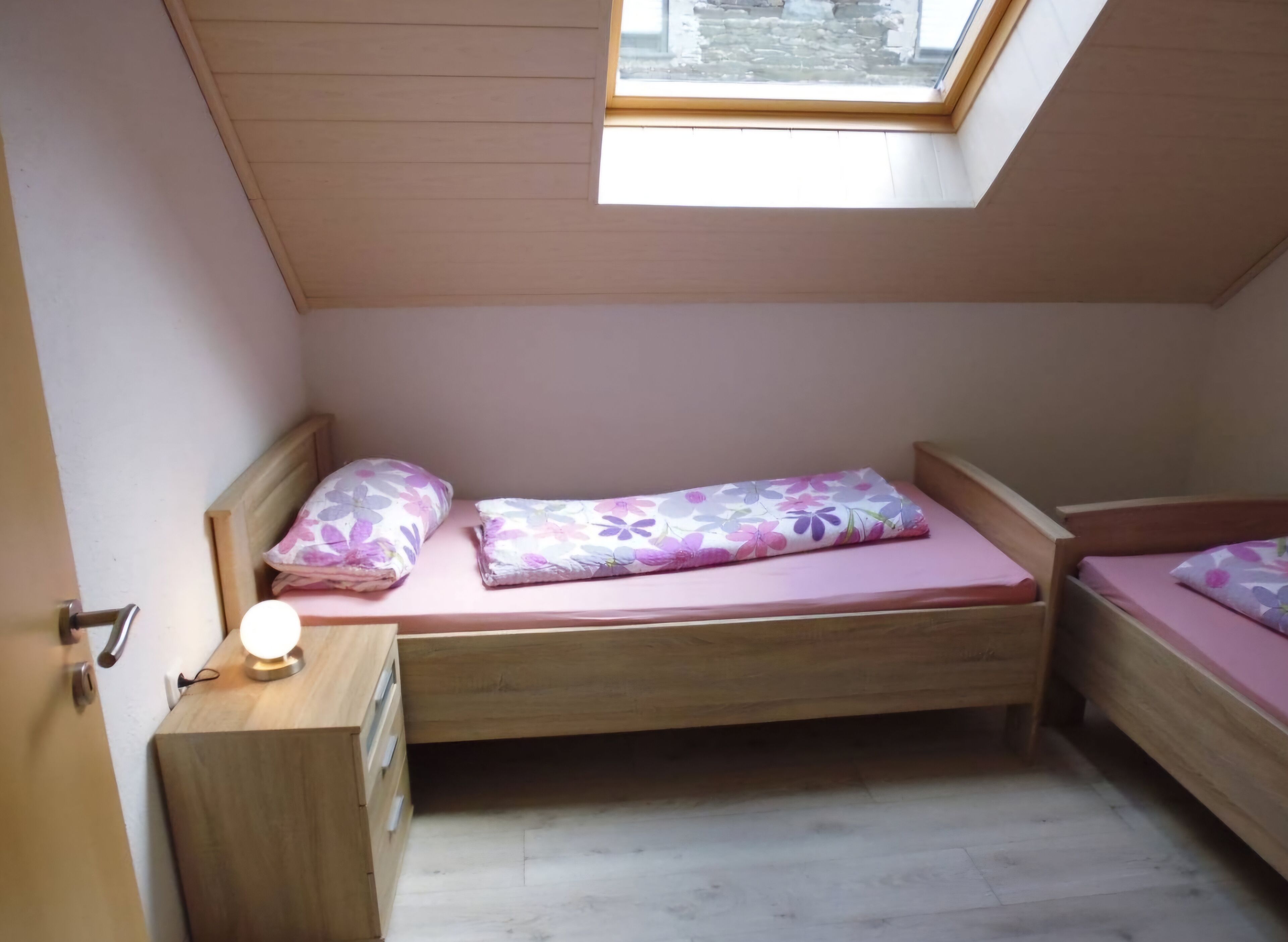 A small, single-room apartment with a sloped ceiling and a skylight. It includes a bed with floral bedding, a small wooden desk with drawers, and a nightstand with a lamp.