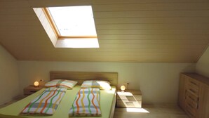 Une petite chambre à coucher accueillante, avec un plafond en pente et un puits de lumière. Elle est équipée d'un lit avec une literie à rayures, de lampes de chevet et d'une armoire en bois.
