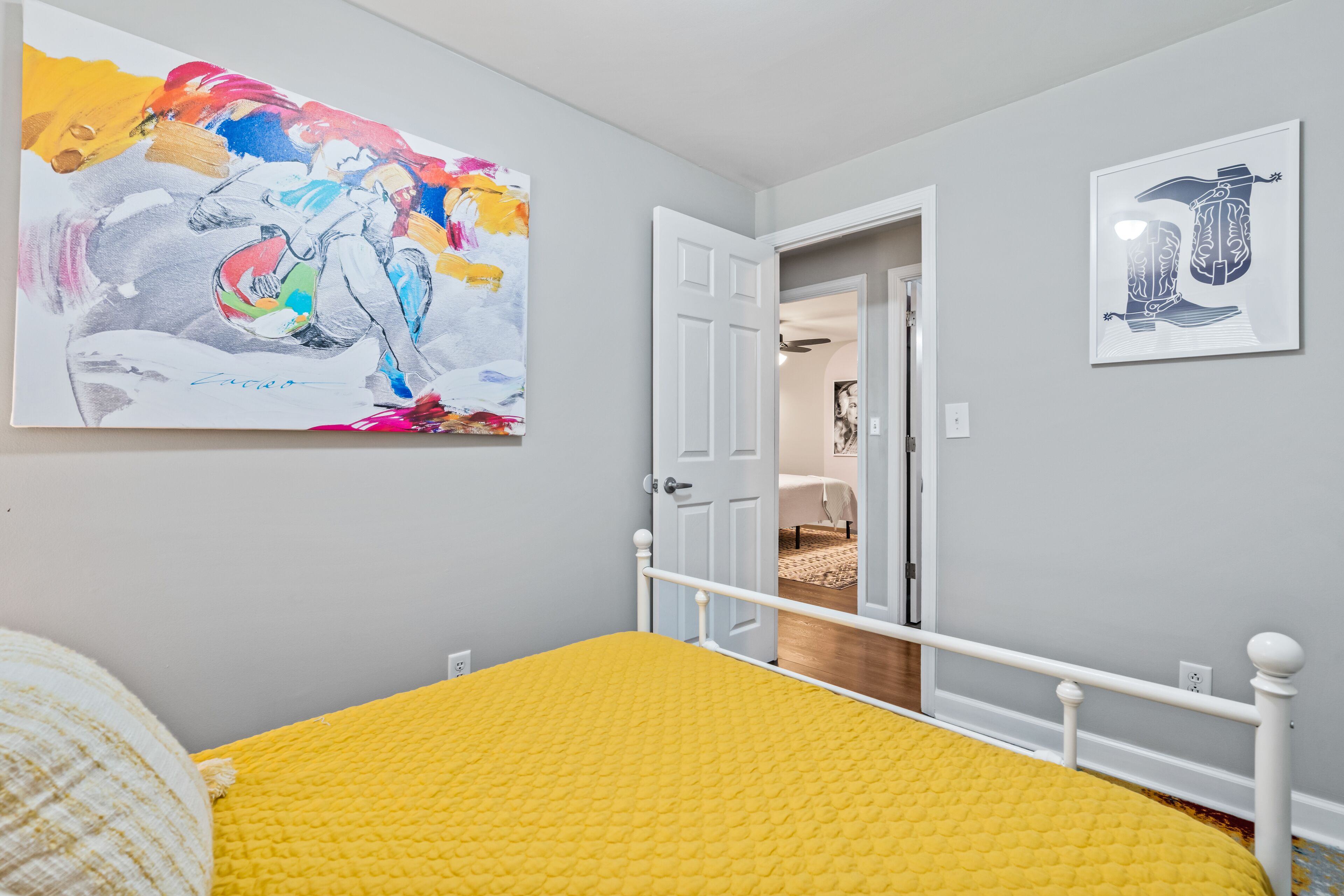 A bedroom with a yellow bedspread, a white bed frame, a colorful abstract painting, and a framed artwork on the wall.