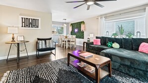 Un salon moderne avec un parquet en bois foncé, un tapis noir et blanc, un canapé vert foncé agrémenté de coussins roses, une table basse en bois et un ventilateur de plafond.