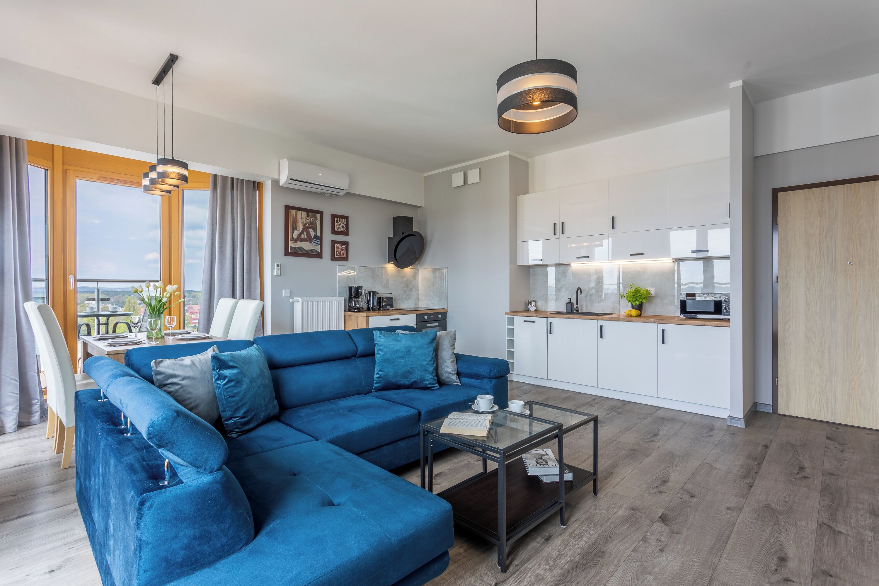 Una sala de estar moderna con un sofá seccional azul, una mesa de centro de vidrio y un comedor con una mesa y sillas blancas.