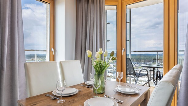 Una mesa de comedor preparada para cuatro, con un jarrón de flores blancas, vistas a la ciudad a través de amplias ventanas y un balcón con mobiliario exterior.