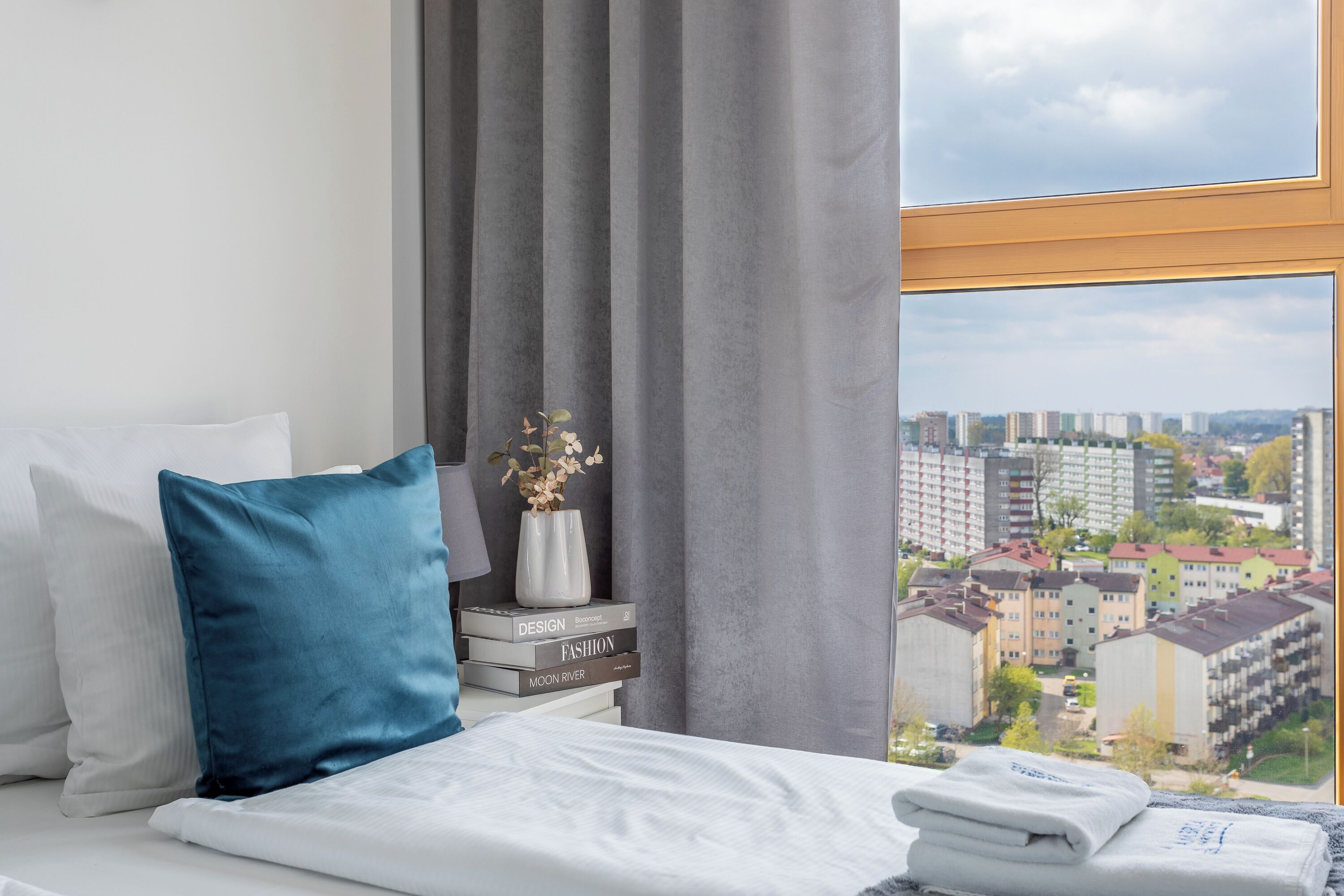 Hotellihuone, jossa on sänky, tyynyjä, pino kirjoja, maljakko kukkineen ja ikkunasta avautuva näkymä kaupunkimaisemaan.