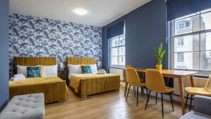 Habitación de hotel con dos camas, una mesa de comedor con sillas, un pequeño banco y un ventanal con vistas a edificios.