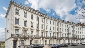 A row of white, multi-story townhouses with uniform architecture, a clear sky, and parked cars along the street. - BRAND NEW The Palms: Luxury 4-6ppl Apartment (Greater London)
