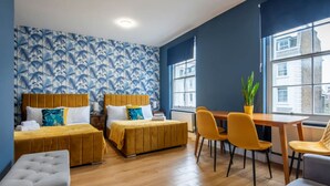 A hotel room with two beds, a dining table with chairs, a small table with a plant, and a window with a view of buildings. - BRAND NEW The Palms: Luxury 4-6ppl Apartment (Greater London)