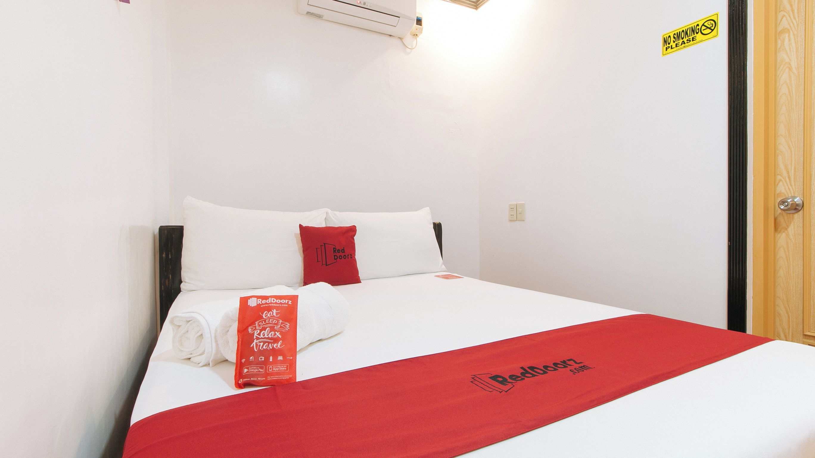 Una cama bien hecha con ropa de cama blanca y roja, una toalla con la marca RedDoorz y un cartel de 'No Fumar' en la pared.