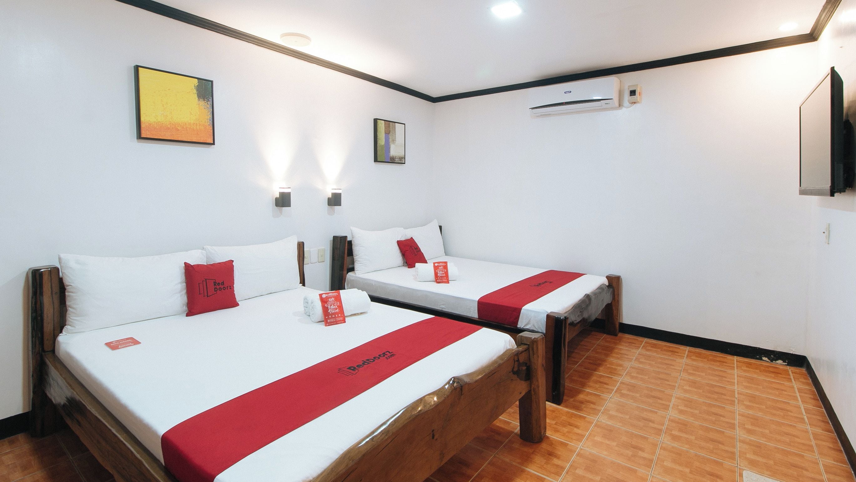 A hotel room with two beds, each with a red bedspread and pillows, a mounted air conditioner, and artwork on the walls.