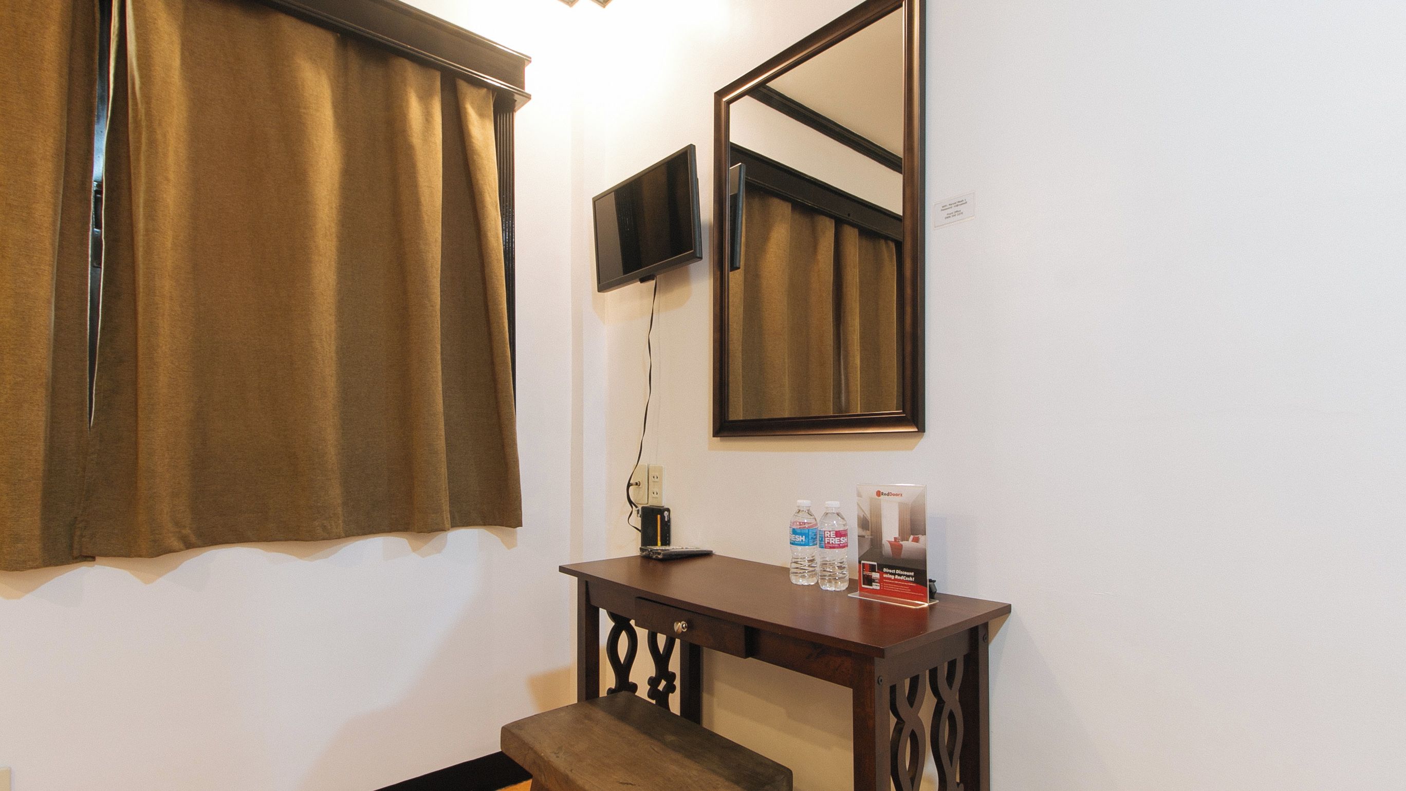 A hotel room with a wooden console table, a flat-screen TV mounted on the wall, a large mirror, and a window with brown curtains.