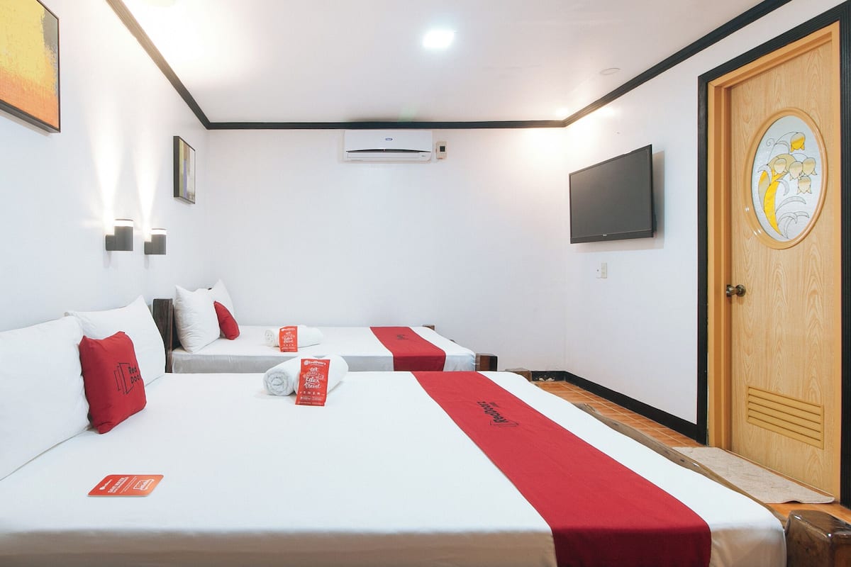 Une chambre d’hôtel avec deux lits, chacun agrémenté de draps blancs et de touches de couleur rouge, un téléviseur à écran plat fixé au mur et une porte en bois avec un panneau de verre décoratif.