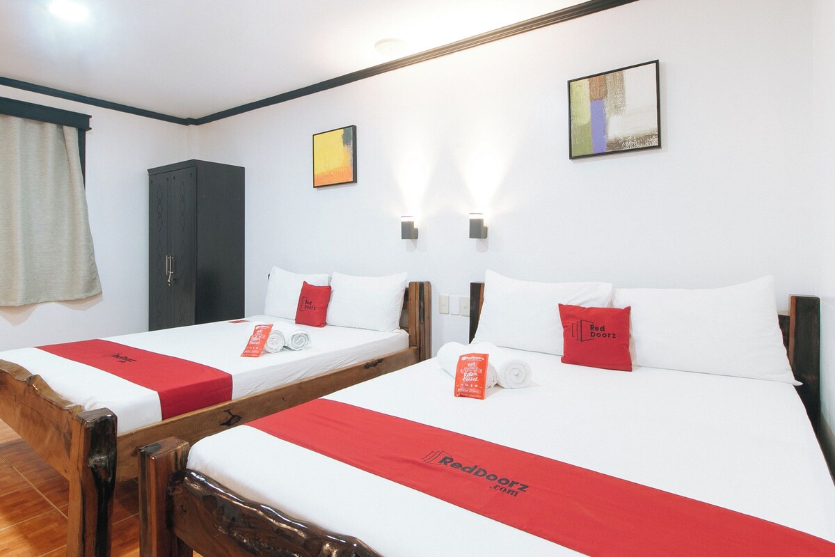 Ein Hotelzimmer mit zwei Einzelbetten, jeweils mit roter Bettdecke und Kissen, einem Holzschrank, einem Fenster mit Vorhängen und einer wandmontierten Lampe.