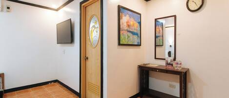 A hallway with a wooden door, a flat-screen TV, a painting, a mirror, and a console table with bottled water.
