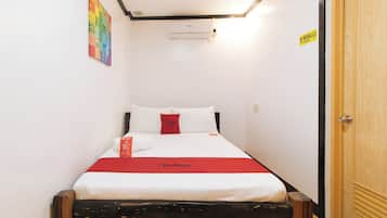 A small, neatly made bed with white and red bedding, a red pillow, and a Red Doorz branded pillow. There is a wall-mounted air conditioning unit above the bed, and a door with a "No Smoking" sign on the wall.