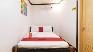 Ein kleines, ordentlich bezogenes Bett mit weißer und roter Bettwäsche, einem roten Kopfkissen und einem Kopfkissen mit dem Branding Red Doorz. Darüber an der Wand ist eine Wand-Klimaanlage angebracht, und an der Wand befindet sich eine Tür mit einem „Nichtraucher“-Schild.