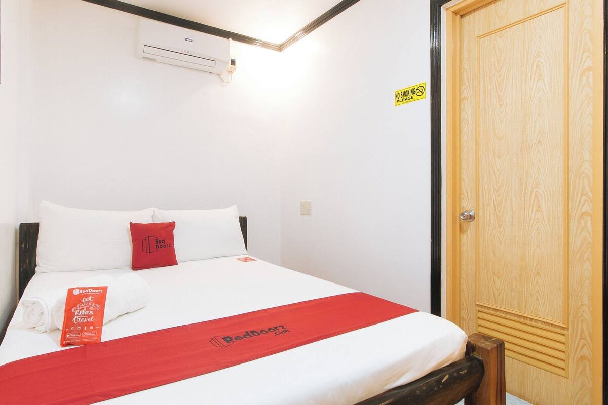Une chambre d’hôtel avec un lit, une couvre-lit rouge et blanche, un oreiller rouge, un climatiseur fixé au mur, une porte en bois et un panneau « Interdiction de fumer ».