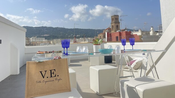 A rooftop terrace with a table set for a meal, a sign reading "V.E. Villa Experiences Puglia", and a view of a church and hills in the background. - Casa Matilda in Fasano (Fasano)