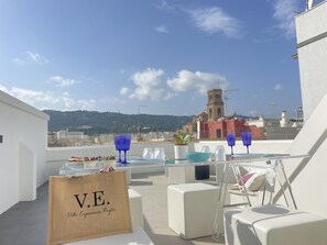 Eine Dachterrasse mit gedecktem Tisch, einem Schild mit der Aufschrift "V.E. Villa Experiences Puglia" und Blick auf eine Kirche und Hügel im Hintergrund.
