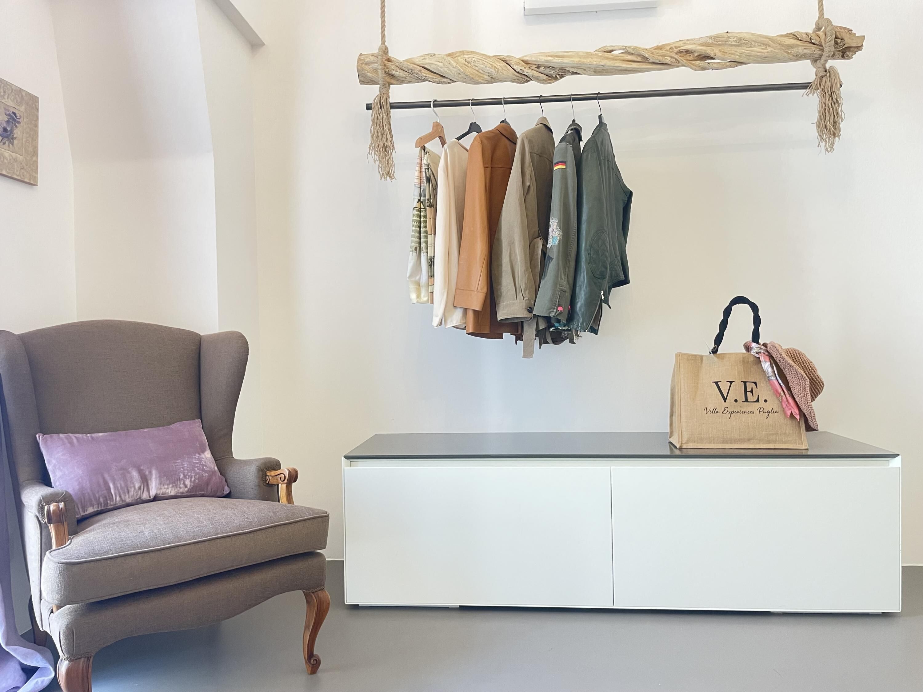 A room with a grey armchair, a white cabinet, and a clothing rack with various garments.