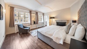 A bedroom with a large bed, a chair, a fireplace, and a view of buildings through the window. - The Chesham Residences (London)
