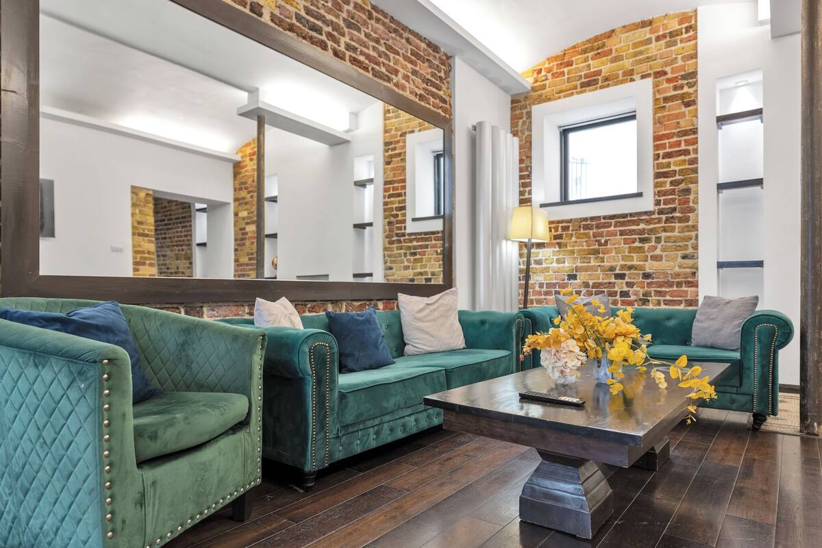 A modern living room with green sofas, a coffee table, and a large mirror reflecting the brick wall.