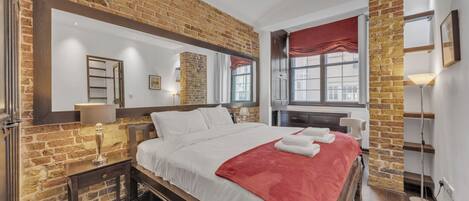 A modern bedroom with a brick wall, a large bed with white bedding and a red throw, a nightstand with a lamp, and a window with a red curtain.