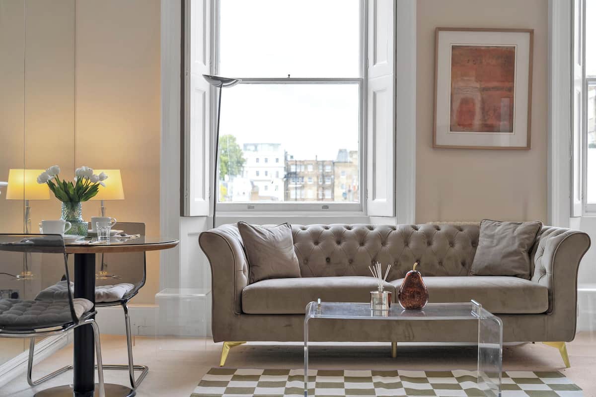 A modern living room with a beige sofa, a glass coffee table, and a view of buildings through large windows.