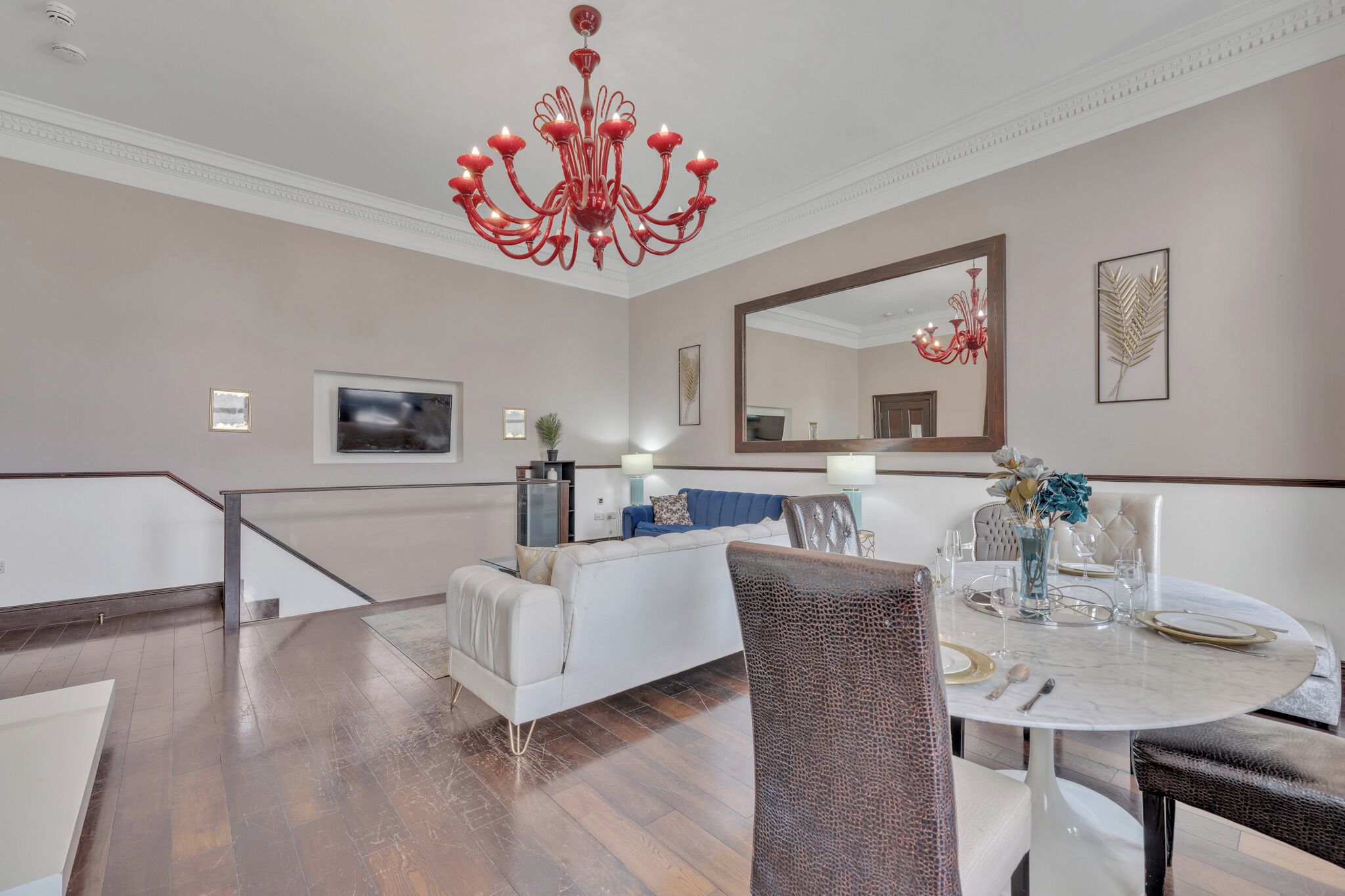 A dining area with a round table set for four, a white sofa, a chandelier, and a large mirror on the wall.