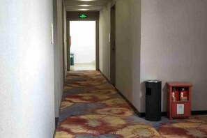 Un couloir d'hôtel avec un sol recouvert de moquette, une poubelle et un petit meuble sur lequel sont posés divers objets.