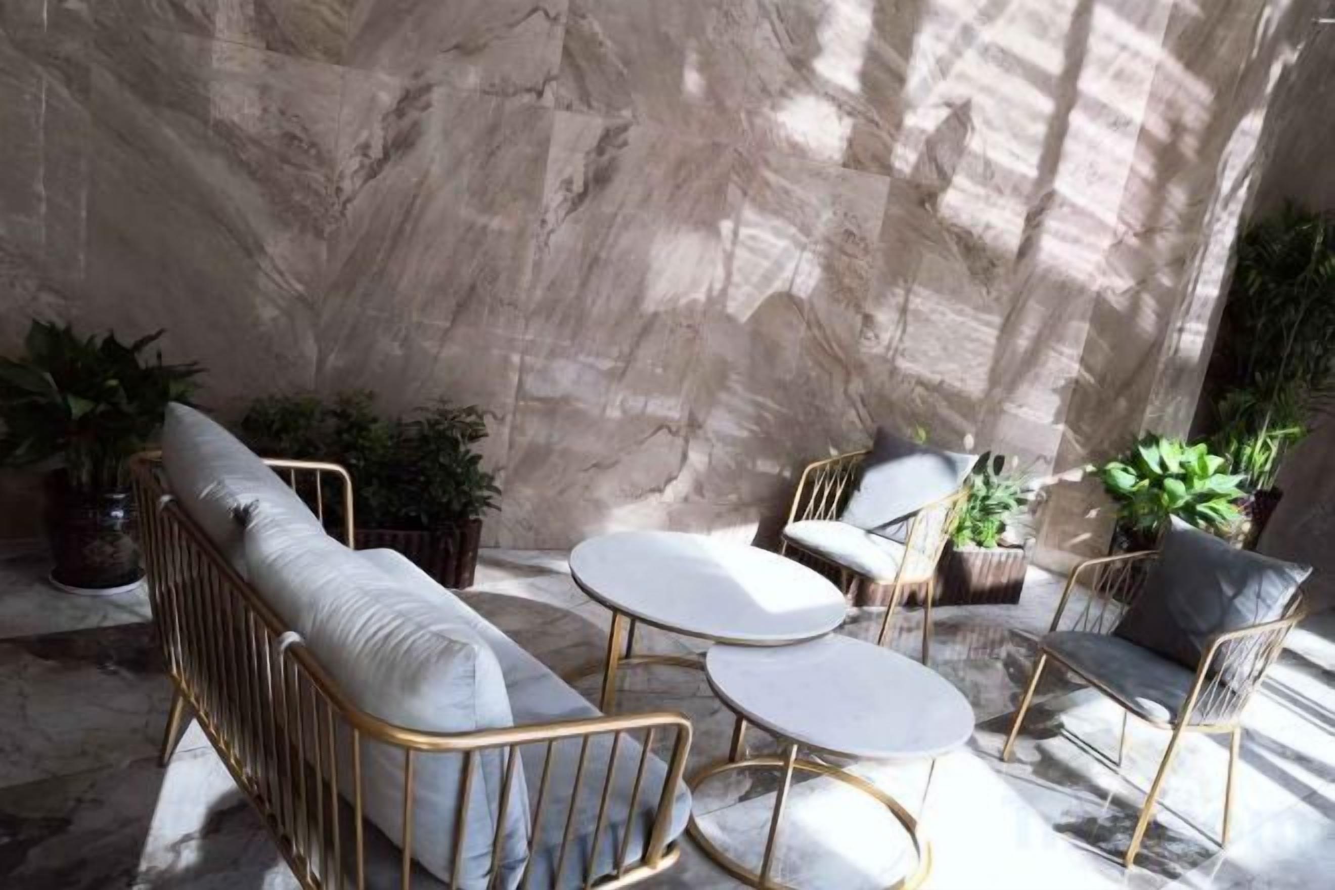 A sunlit patio with a sofa, two round tables, and chairs, surrounded by potted plants.