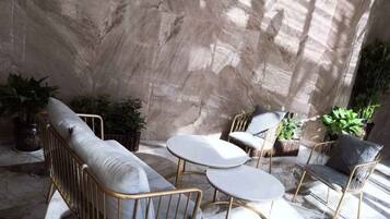 A sunlit patio with a sofa, two round tables, and chairs, surrounded by potted plants.