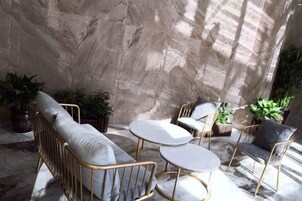 A sunlit patio with a sofa, two round tables, and chairs, surrounded by potted plants.