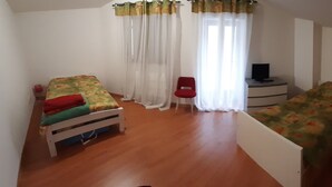 Una habitación pequeña y luminosa con una cama, una silla, una mesita con televisor y un ventanal con cortinas.